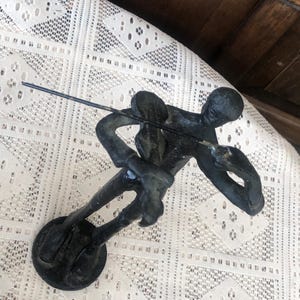 May include: A dark bronze sculpture of a person fishing, with a rod extended. The figure is in a dynamic pose, suggesting movement. The sculpture is mounted on a circular base. The background is a white lace tablecloth.