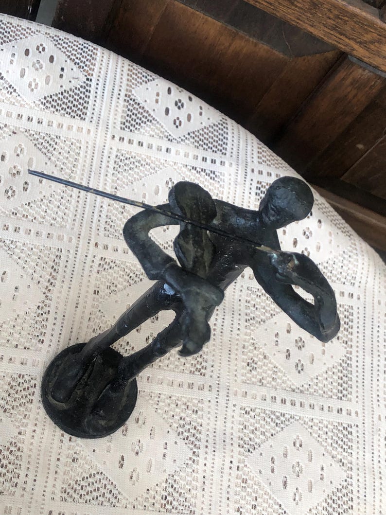 May include: A dark bronze sculpture of a person fishing, with a rod extended. The figure is in a dynamic pose, suggesting movement. The sculpture is mounted on a circular base. The background is a white lace tablecloth.