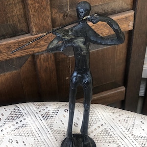 May include: A dark bronze sculpture of a person playing a violin. The figure is stylized with elongated limbs and a round base. The violin bow is held in the right hand, and the instrument is held in the left hand. The sculpture is placed on a white lace surface.