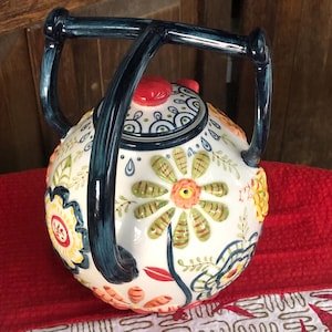 May include: A white ceramic teapot with a floral design featuring orange, yellow, and green flowers. The teapot has a dark blue handle and lid, and is sitting on a red surface.