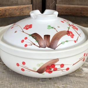 May include: A white ceramic lidded pot featuring a hand-painted design of brown branches with small red berries and light green leaves. The pot has two side handles and a matching lid with a central knob, decorated with the same floral motif.