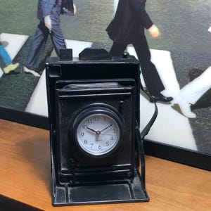 May include: A black metal camera-shaped clock with a white clock face. The clock has a quartz movement and a black leather strap. The clock is sitting on a wooden surface. The background features a framed image of people walking on a crosswalk.