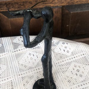 May include: A black metal sculpture of a figure holding an object aloft. The figure stands on a circular base. The sculpture has a rough, textured appearance and is set against a lace tablecloth and wooden background.