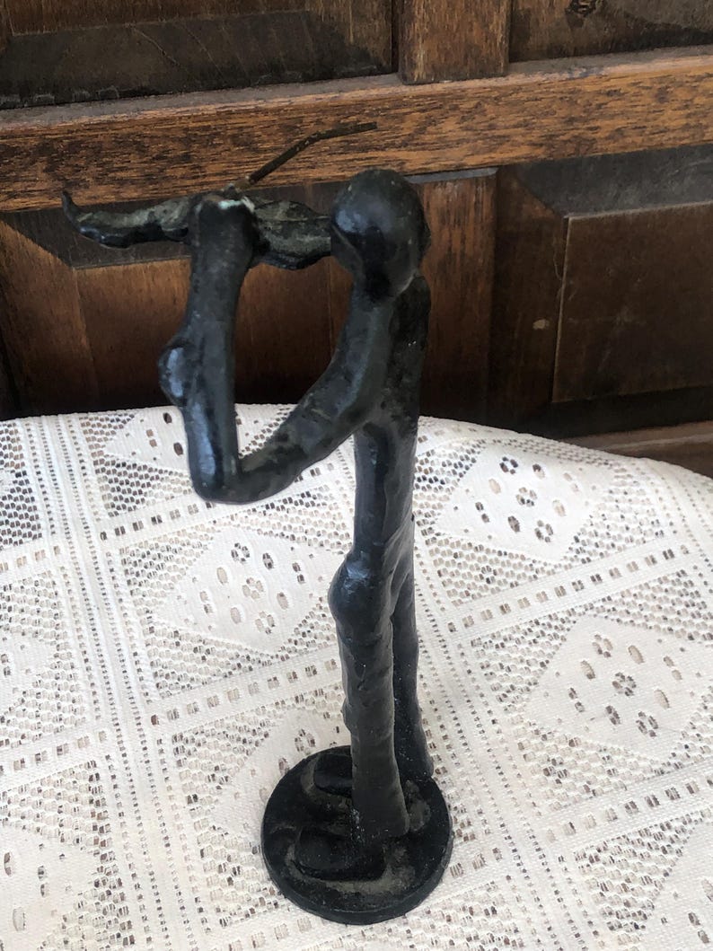 May include: A black metal sculpture of a figure holding an object aloft. The figure stands on a circular base. The sculpture has a rough, textured appearance and is set against a lace tablecloth and wooden background.