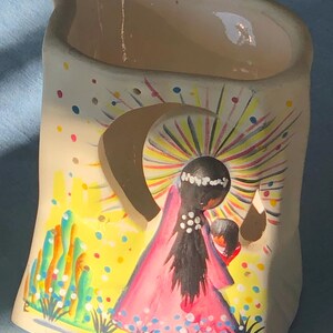 May include: A ceramic candle holder with a hand-painted design. The artwork depicts a figure in a pink robe with a halo, holding a child. The background is yellow with colorful dots and rays. The name "DeGrazia" is written on the bottom.