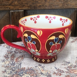May include: A red ceramic mug featuring a dark brown base with small yellow dots, a handle with yellow accents, and a repeating pattern of brown oval shapes containing blue and red floral motifs. The interior rim has small red floral icons.