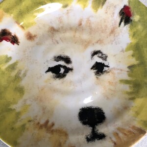May include: A ceramic plate with a hand-painted dog face design. The dog has black eyes and nose, with tan and white fur. The plate is bordered with yellow and green.