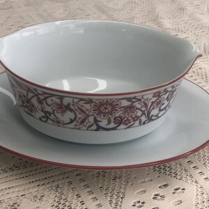 May include: A white porcelain gravy boat with a floral design in shades of red, pink, and blue. The boat is sitting on a matching saucer.