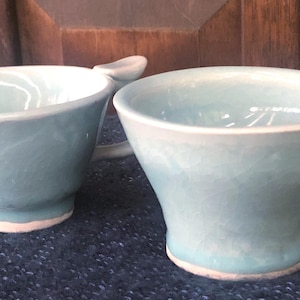 May include: Two handmade ceramic mugs in a light blue color. The mugs have a unique handle design and a white rim. The mugs are sitting on a dark blue surface.