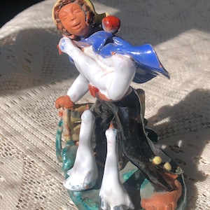 May include: A ceramic figurine of a person with brown skin, wearing a blue and yellow hat, a blue cloak, and black pants, holding a white goose in their arms. The person is walking with two white geese following behind them. The figurine is painted with details and has a green base.