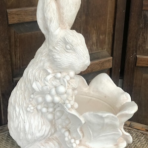 May include: A white rabbit-shaped planter with a large, open flower-like bowl. The rabbit is detailed with textured fur and holds a cluster of grapes. The planter is made of a light-colored material, suitable for indoor or outdoor use.