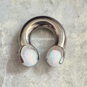 Op de afbeelding: Een zilverkleurig, hoefijzervormig lichaamssieraad met twee witte, opaalachtige uiteinden. De sieraden hebben een gladde, gepolijste afwerking en zijn geplaatst op een lichtgrijze, marmerachtige ondergrond. De tekst "@midsparkgems" is zichtbaar.