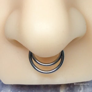 May include: A close-up of a double hoop nose ring made of black metal. The ring is worn in a person's nose.