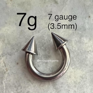 May include: A silver-colored, horseshoe-shaped piercing with two conical, pointed ends. The text "7g" and "7 gauge (3.5mm)" is displayed above the piercing. The piercing is designed for body modification.