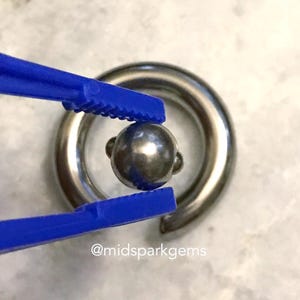 Pode incluir: Um anel de contas cativas prateado com uma conta a condizer presa por pinças azuis. A joia é exibida numa superfície de cor clara. A conta tem um diâmetro de aproximadamente 0,6 cm.