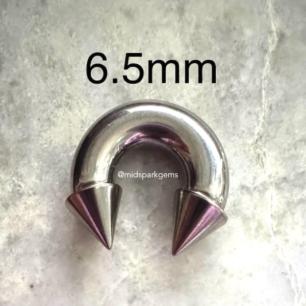 Puede incluir: Un piercing plateado en forma de herradura con dos extremos cónicos con puntas moradas. El texto "6.5mm" está encima del piercing, y el nombre de usuario de Instagram "@midsparkgems" debajo.