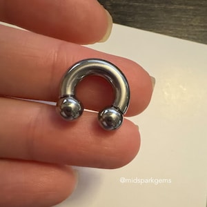 以下が含まれることがあります： 2 つのボールエンドが付いたシルバーの円形バーベル。バーベルは手で持たれています。