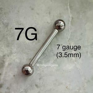May include: A silver-colored barbell-style body piercing jewelry. The jewelry has a straight bar with a ball on each end. The text "7G" and "7 gauge (3.5mm)" is visible in the image. The jewelry is displayed on a light-colored, textured surface.