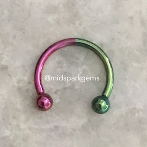 May include: A circular horseshoe-shaped earring with a pink and green iridescent finish. The earring is made of metal and has a smooth surface.