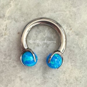 Puede incluir: Una barra de herradura de plata con extremos de ópalo azul. La barra es un tipo de joyería corporal.