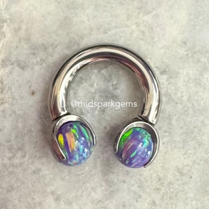 Puede incluir: Una barra de herradura de plata con extremos de ópalo iridiscente morado y verde. La barra es un tipo de joyería corporal.