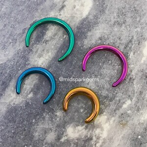 May include: Four iridescent half-circle rings in different colors: teal, purple, blue, and gold. The rings are on a gray marble surface. @midsparkgems