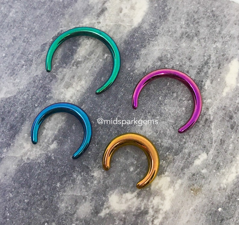 May include: Four iridescent half-circle rings in different colors: teal, purple, blue, and gold. The rings are on a gray marble surface. @midsparkgems