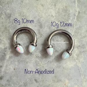 May include: Two silver circular barbell earrings with white opal ends. The earring on the left is 8 gauge and 10mm in diameter. The earring on the right is 10 gauge and 12mm in diameter. The text "Non-Anodized" is below the earrings.
