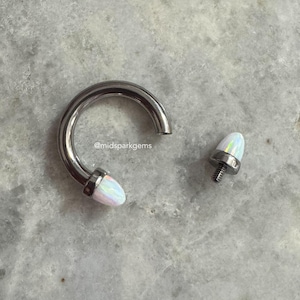May include: A silver septum ring with a white opal gem on the end. The ring is open and the gem is separate.