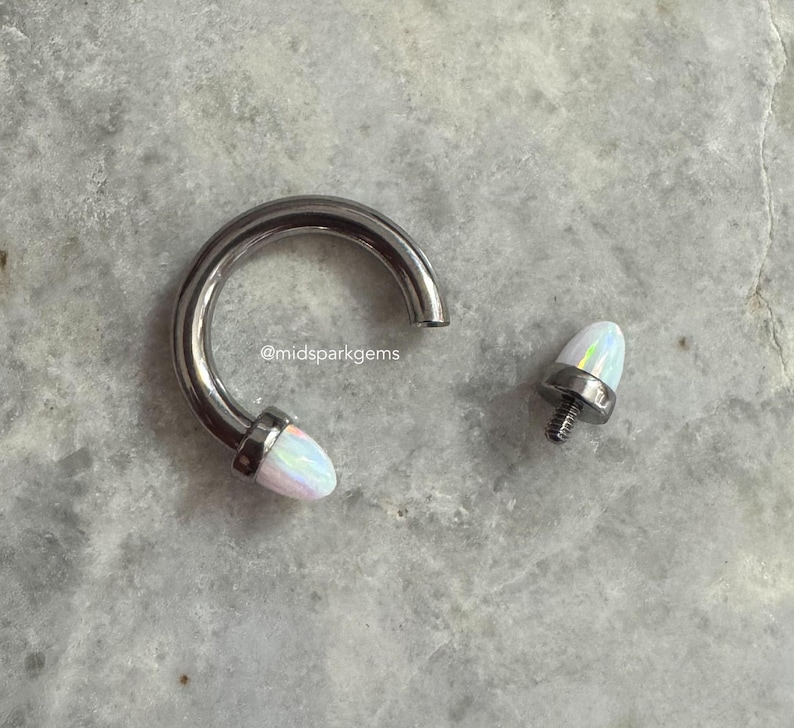 May include: A silver septum ring with a white opal gem on the end. The ring is open and the gem is separate.