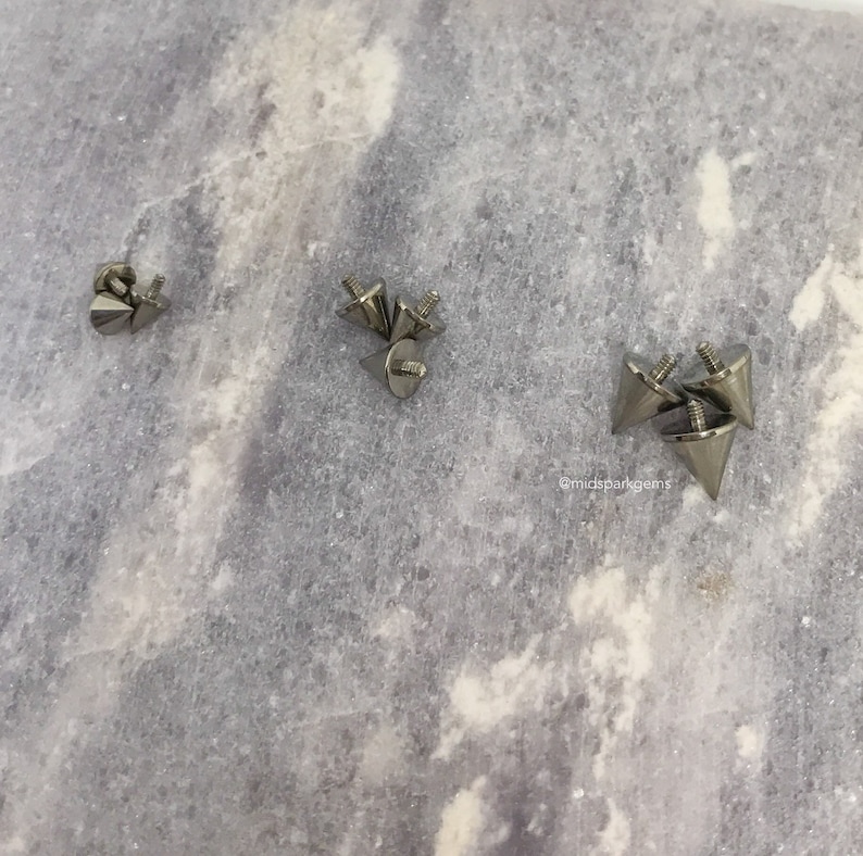 May include: Several silver-colored, cone-shaped metal studs with screw-on bases are scattered across a light gray surface. The studs have a pointed tip and a threaded base for attachment. The image is well-lit, highlighting the metallic sheen of the studs.