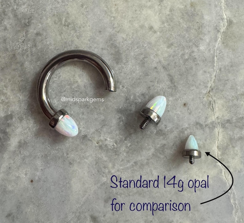 May include: A close-up of three opal body jewelry pieces. The largest piece is a silver hoop with a white opal on the end. The other two pieces are smaller white opal studs. The text "Standard 14g opal for comparison" is written below the image.