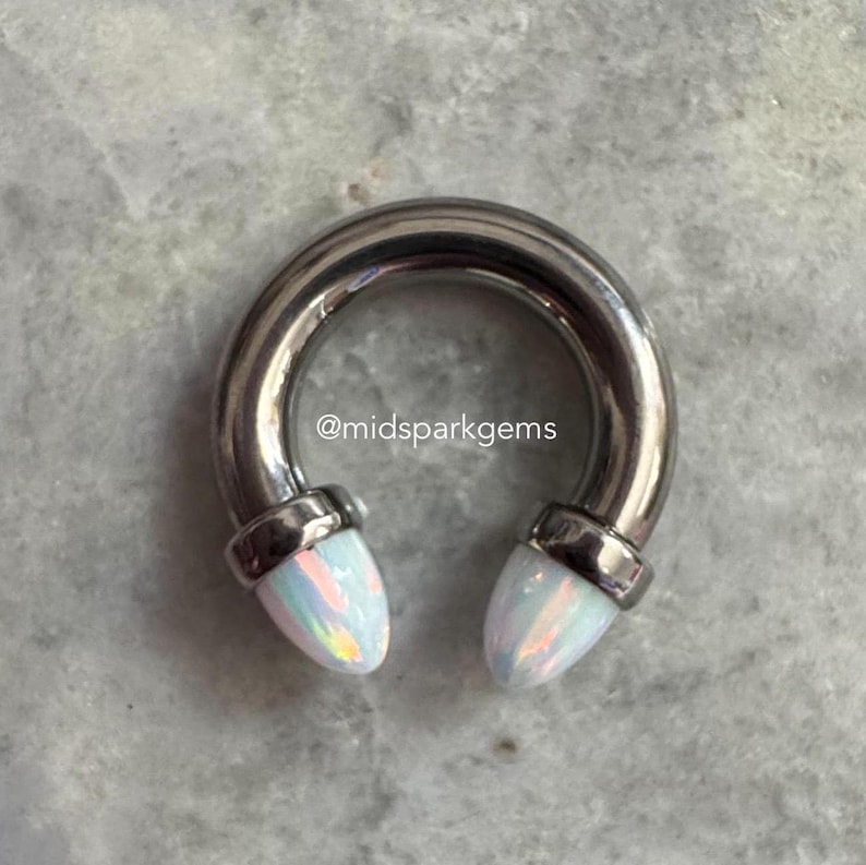 May include: A silver horseshoe-shaped barbell with opal-like ends. The ends are a light iridescent white with hints of pink and blue.