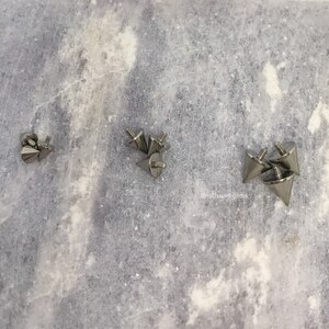 May include: A collection of silver-colored, cone-shaped metal studs with screw-on backs. The studs are arranged on a light gray, textured surface. The studs have a pointed tip and a flat base.