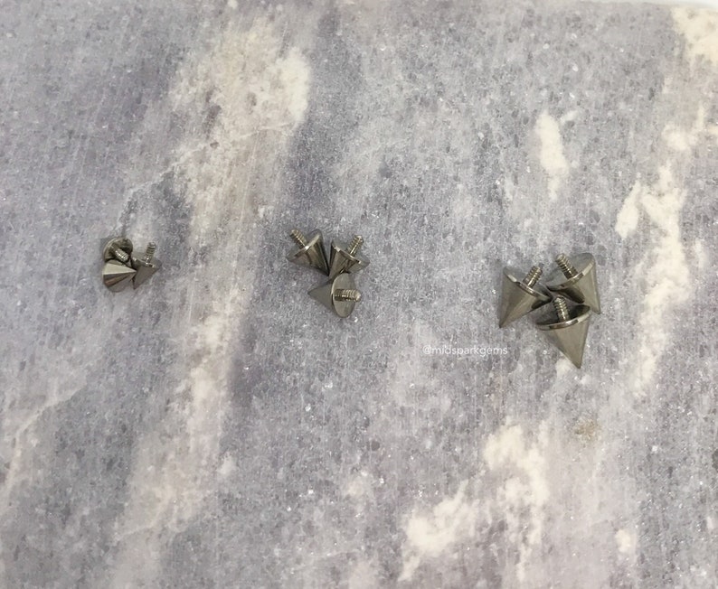 May include: A collection of silver-colored, cone-shaped metal studs with screw-on backs. The studs are arranged on a light gray, textured surface. The studs have a pointed tip and a flat base.