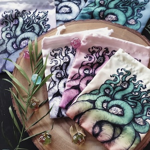 Könnte beinhalten: Fünf bunte Kordelbeutel mit einem Motiv einer grünen, tentakelartigen Kreatur mit aufgedruckten Würfeln. Die Beutel sind auf einer Holzoberfläche mit verstreuten Würfeln angeordnet.