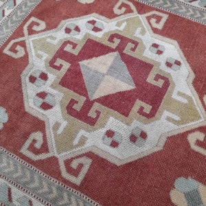 May include: A red, white, and beige patterned rug with a geometric design. The rug has a border of white and beige.