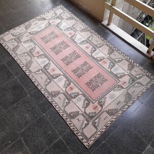 May include: A rectangular rug with a central pink panel and geometric patterns. The rug has a cream border with grey and pink designs. It is placed on a dark grey tiled floor. The rug's design is traditional.