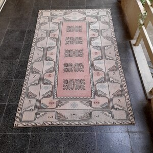 May include: A rectangular rug with a central pink panel and a patterned border. The rug features geometric designs in black, white, and grey, creating a vintage aesthetic. The rug is placed on a dark tiled floor.
