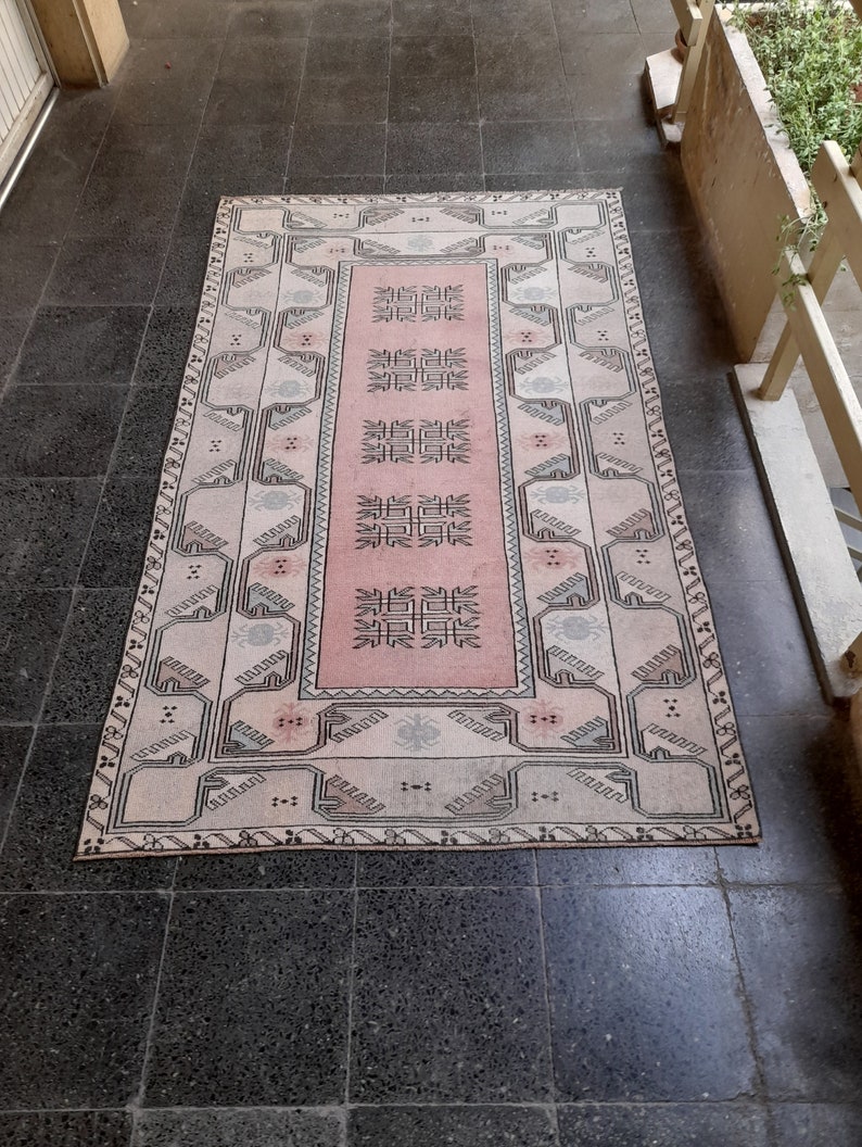 May include: A rectangular rug with a central pink panel and a patterned border. The rug features geometric designs in black, white, and grey, creating a vintage aesthetic. The rug is placed on a dark tiled floor.