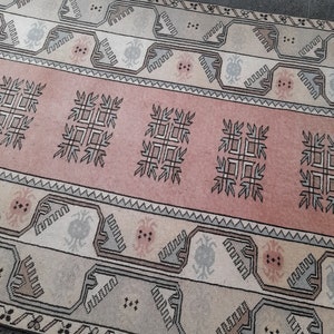 May include: A rectangular rug with a central pink band featuring geometric patterns. The rug has cream and grey borders with repeating designs. The rug's overall design is symmetrical and traditional, suitable for home decor.