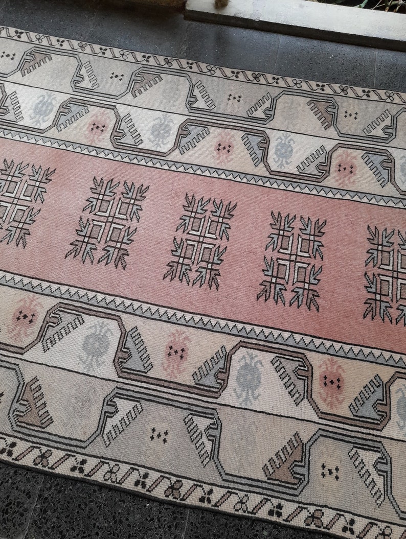 May include: A rectangular rug with a central pink band featuring geometric patterns. The rug has cream and grey borders with repeating designs. The rug's overall design is symmetrical and traditional, suitable for home decor.