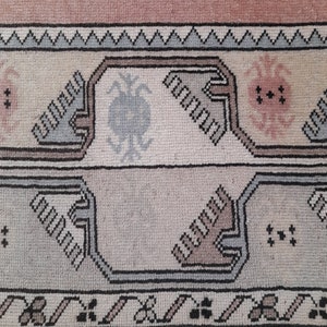 May include: Close-up of a vintage rug with a geometric pattern. The rug features a colour palette of pink, beige, grey, and black. The design includes repeating shapes and floral motifs, creating a textured appearance.