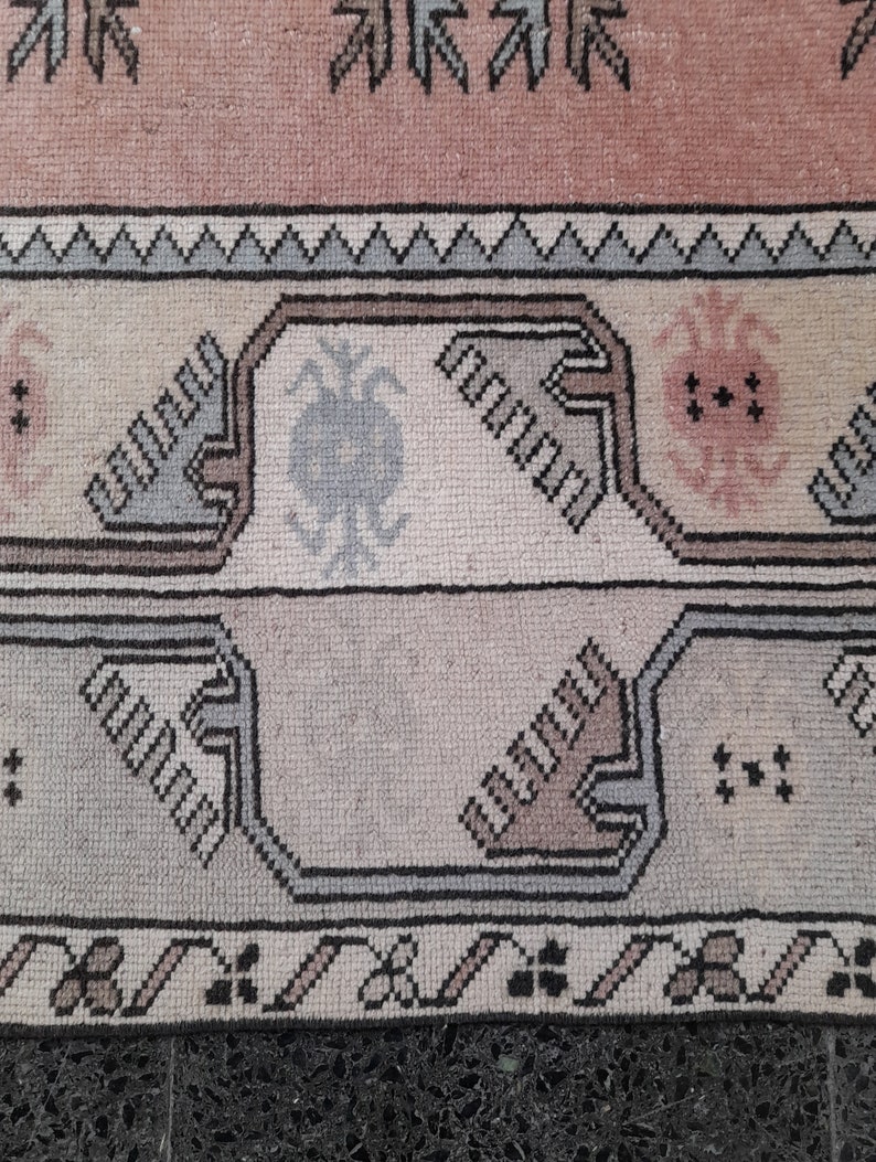 May include: Close-up of a vintage rug with a geometric pattern. The rug features a colour palette of pink, beige, grey, and black. The design includes repeating shapes and floral motifs, creating a textured appearance.