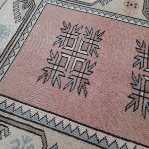 May include: Close-up of a vintage rug with a central pink field and detailed geometric patterns. The rug has a border with repeating shapes in shades of grey, blue, and beige. The central design includes stylized floral and geometric motifs.