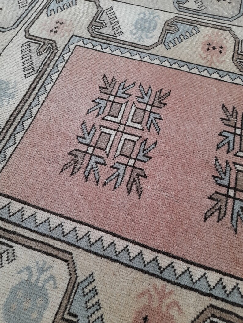 May include: Close-up of a vintage rug with a central pink field and detailed geometric patterns. The rug has a border with repeating shapes in shades of grey, blue, and beige. The central design includes stylized floral and geometric motifs.