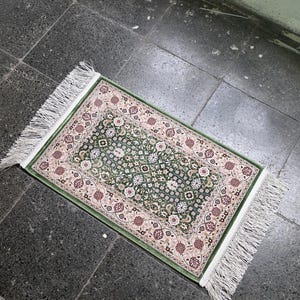 May include: A rectangular green rug with a floral pattern and white fringe. The rug features a central field of green with white and burgundy floral designs, framed by a beige border with similar patterns. The rug is approximately 24 inches long and 16 inches wide.