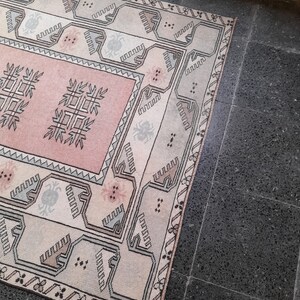 May include: A rectangular rug with a central pink panel featuring geometric designs. The rug has a cream-coloured border with repeating patterns in black, grey, and pink. The rug is placed on a dark grey tiled floor.