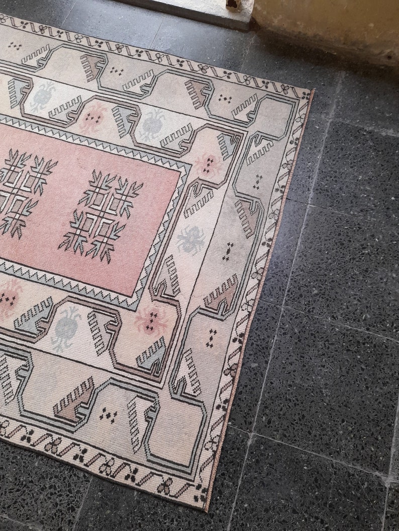 May include: A rectangular rug with a central pink panel featuring geometric designs. The rug has a cream-coloured border with repeating patterns in black, grey, and pink. The rug is placed on a dark grey tiled floor.