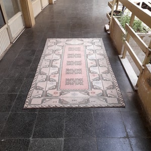 May include: A rectangular rug with a central pink panel and a cream border. The rug has a geometric pattern in pink, cream, and grey. It's placed on a dark grey tiled floor, with a building's exterior visible in the background. The rug's design is intricate.